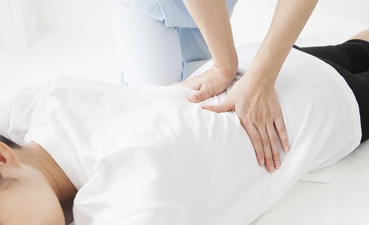 사람이 하얀색 옷을 입고 엎드려 누워 있으며, 다른 사람이 그 위에 손을 올려 마사지를 하고 있습니다. 마사지하는 사람은 하늘색 옷을 입고 있습니다. 배경은 밝고 깨끗한 분위기입니다.