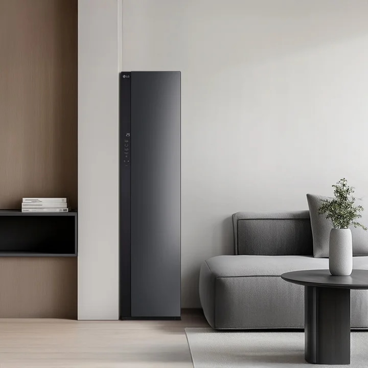 LG 스타일러 제품이 방의 왼쪽에 배치되어 있으며, 슬림한 디자인과 어두운 색상의 전면 패널이 특징입니다. 스타일러 옆에는 회색 소파가 놓여 있고, 소파 위에는 회색 쿠션이 있습니다. 소파 앞에는 작은 원형 테이블이 있으며, 테이블 위에는 흰색 꽃병과 꽃이 놓여 있습니다. 방의 오른쪽에는 큰 창문이 있어 자연광이 들어오고 있습니다.