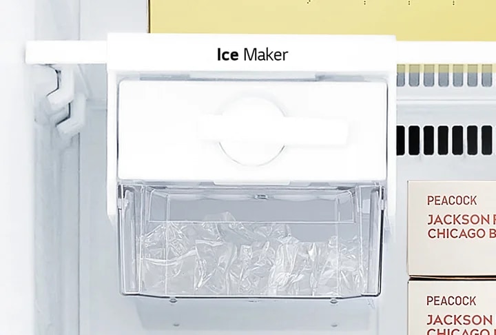 냉장고 내부의 아이스 메이커가 보입니다. 아이스 메이커 위에는 Ice Maker라는 텍스트가 적혀 있습니다. 아이스 메이커 아래에는 투명한 용기에 얼음이 담겨 있습니다. PEACOCK JACKSON CHICAGO B라는 텍스트가 적힌 상자가 보입니다.