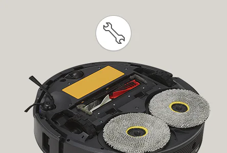로봇 청소기의 하단부가 보이며, 두 개의 회전 브러시와 중앙에 위치한 흡입구가 눈에 띕니다. 흡입구 옆에는 노란색 커버가 덮여 있습니다. 이미지 상단에는 렌치 모양의 아이콘이 있습니다. 배경은 회색입니다.